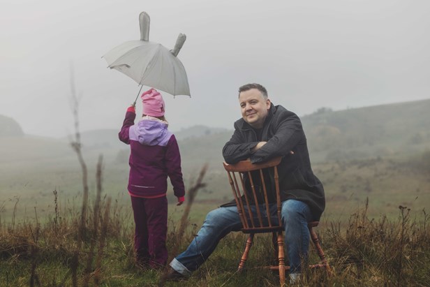Conny Palmkvist sitter på en stol utomhus, bredvid honom står ett barn med ryggen mot kameran och ett uppfällt paraply.