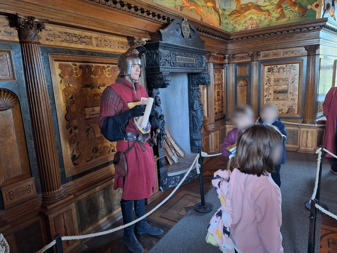 En museiplats i slottet med riddare i röd rustning som håller en skriftrulle, omgiven av utsmyckade träpaneler och en publik av barn.