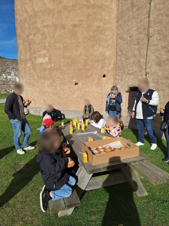 Det är en solig dag, en grupp med deltagare bestående av barn och vuxna, njuter av snacks och drycker runt ett picknickbord med slottet i bakgrunden.