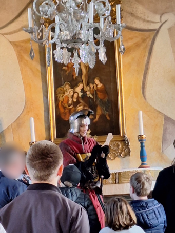 En person i historisk rustning läser ett dokument för en grupp i ett rum på slottet. En ljuskrona och religiösa målningar skapar en högtidlig atmosfär.